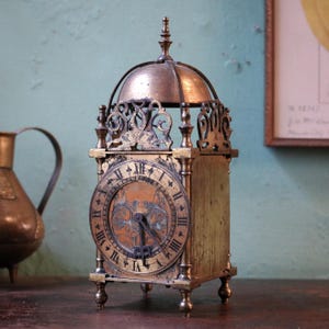 May include: A metallic lantern-style clock featuring a dome top, a circular face with black Roman numerals, and intricate filigree detailing. The clock is supported by four small feet and displays a weathered, aged finish with dark metal accents.