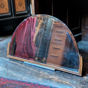 May include: A semi-circular wall mirror featuring a beveled glass edge and a wooden frame with a stepped, tiered design at the base corners. The mirror reflects a wooden chest of drawers and red fabric, with a silver-toned metal support bar.