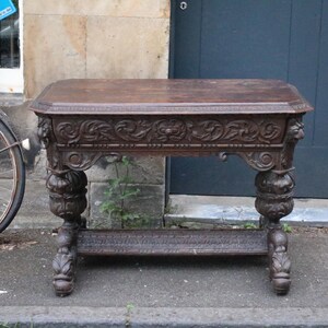 May include: An antique dark brown wooden table with intricate carvings. The table has floral and lion head designs on the apron and legs. It is rectangular with a beveled edge and stands on four elaborately carved legs. The table measures approximately 120 cm in length.