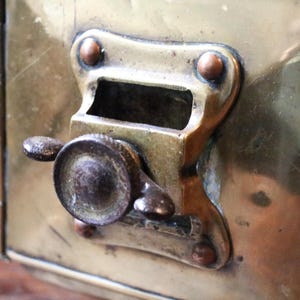 May include: Close-up of a vintage brass box with a hinged lid and a locking mechanism. The box has a tarnished finish, showing signs of age. The locking mechanism features a small handle and a rectangular opening. The box is likely used for storage.