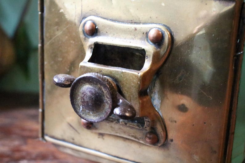 May include: Close-up of a vintage brass box with a hinged lid and a locking mechanism. The box has a tarnished finish, showing signs of age. The locking mechanism features a small handle and a rectangular opening. The box is likely used for storage.
