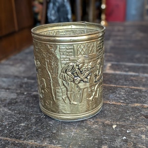 Puede incluir: Un porta lápices de metal dorado con un diseño en relieve de personas en una escena de taberna.