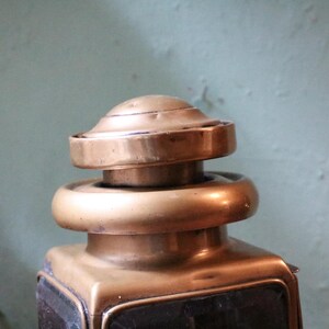 May include: A close-up of an antique brass lantern. The lantern has a square base, a rounded top, and a decorative finial. The brass has a warm, aged patina, indicating its age. The background is a muted green colour.