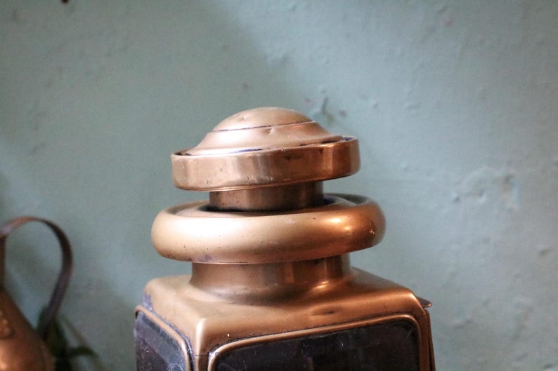 May include: A close-up of an antique brass lantern. The lantern has a square base, a rounded top, and a decorative finial. The brass has a warm, aged patina, indicating its age. The background is a muted green colour.