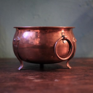 May include: A copper cauldron with a rounded body, three curved legs, and a large ring handle. The cauldron has a polished finish, reflecting the surrounding light. Decorative rivets and accents are visible on the sides. The cauldron is a vintage piece.