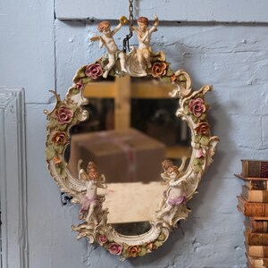 May include: Oval-shaped decorative mirror with a cream-colored frame adorned with cherub figurines and pink and orange roses. Two cherubs are at the top, and two are at the bottom. The mirror hangs from a chain.