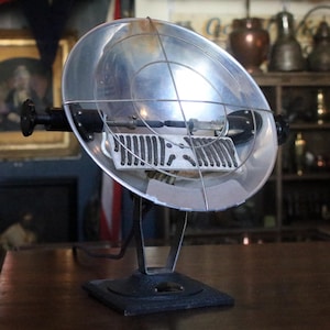 May include: A retro electric heater with a silver reflector and a protective wire guard. The heater has a black base and adjustable controls. The design suggests a vintage or industrial aesthetic, suitable for home decor. The measurements are in centimeters.