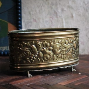 Puede incluir: Un recipiente ovalado de latón antiguo con detalles en relieve de frutas y follaje. El recipiente descansa sobre pequeñas patas en forma de garra. El latón tiene una pátina cálida y envejecida.