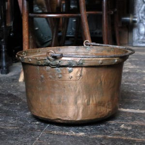 Puede incluir: Un gran caldero de cobre antiguo con un borde remachado y un asa de metal resistente. La olla tiene un cuerpo redondeado y una rica pátina envejecida, lo que sugiere años de uso. El fondo incluye muebles de madera.