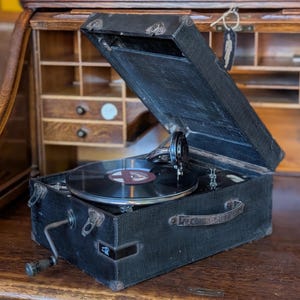 Op de afbeelding: Een zwarte draagbare koffergrammofoon met een getextureerde buitenkant, een metalen zwengel aan de zijkant en een centrale draaitafel met een zwarte vinylplaat. Het deksel staat open en toont de interne toonarm en naaldmechanisme.