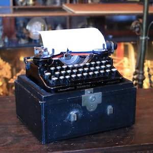 May include: A vintage black Underwood Standard typewriter with a white sheet of paper inserted. The typewriter sits atop a black case with a metal clasp. The keys are white and the machine has a silver carriage.