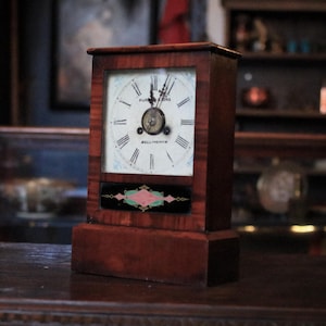 Puede incluir: Un reloj de madera antiguo de forma rectangular. La esfera es blanca con números romanos negros y la inscripción "MURDOCK & SONS BELLINGHAM". El reloj tiene un panel decorativo con un diseño rosa y verde.