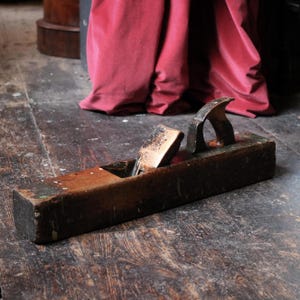 May include: An antique wooden hand plane, showing signs of wear and age. The plane has a dark, weathered wooden body with a metal blade and handle. The wood has a warm, brown tone. The tool is resting on a wooden floor, with a red curtain in the background.