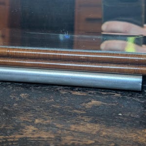 May include: A rectangular mirror featuring a polished wooden frame with a smooth, rounded edge. Below the wooden frame, a cylindrical silver-toned metal bar runs horizontally across the width of the mirror, providing a distinct contrast in texture.