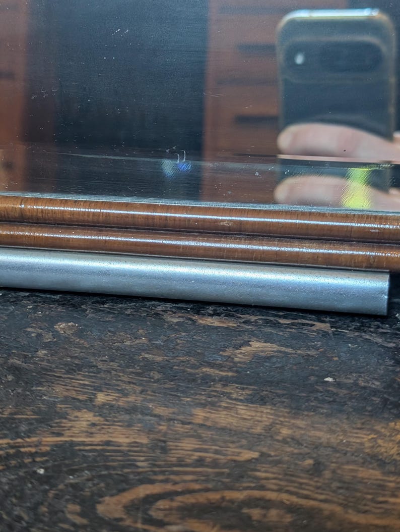 May include: A rectangular mirror featuring a polished wooden frame with a smooth, rounded edge. Below the wooden frame, a cylindrical silver-toned metal bar runs horizontally across the width of the mirror, providing a distinct contrast in texture.