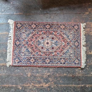 Puede incluir: Una alfombra rectangular con un diseño de medallón central que presenta intrincados patrones florales y geométricos en tonos terracota, azul marino y crema. La alfombra tiene un borde decorativo y flecos de color blanco roto en los extremos.
