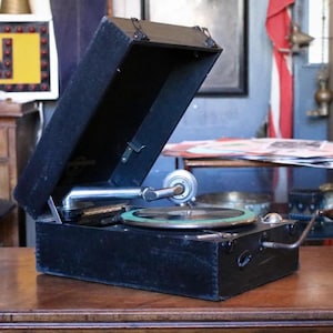May include: An antique black gramophone with the lid open, revealing the turntable and arm. The gramophone sits on a wooden surface, showcasing its vintage design and mechanical components. The turntable has a green border.