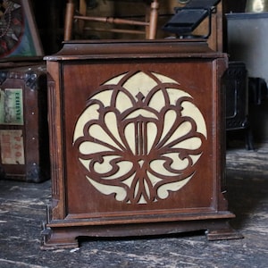 May include: A square, dark brown wooden radio cabinet featuring a large, circular front panel with an intricate, symmetrical cutout design layered over a light cream-coloured fabric speaker grille. The wooden frame has a flat top and base.
