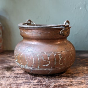 May include: An antique copper pot with a hammered finish and engraved details. The pot has two metal handles and a lid.