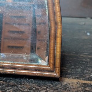 May include: A wooden mirror with a curved frame featuring a light brown wood grain finish. The reflective glass surface shows a partial reflection of a wooden cabinet with multiple drawers, capturing the depth and detail of the wood texture.