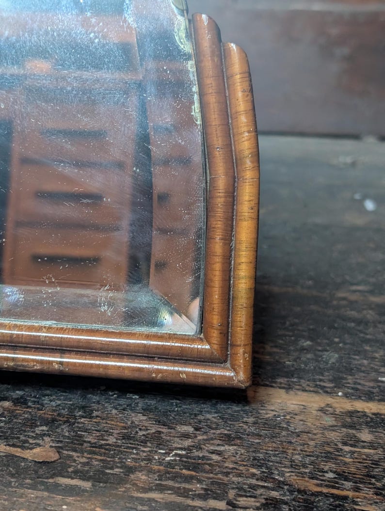May include: A wooden mirror with a curved frame featuring a light brown wood grain finish. The reflective glass surface shows a partial reflection of a wooden cabinet with multiple drawers, capturing the depth and detail of the wood texture.