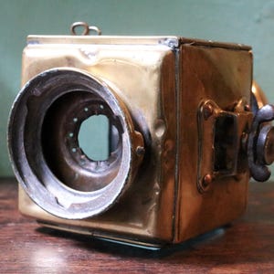 May include: Antique brass camera, square-bodied with a large circular lens. It features a dark metal focusing knob and small rectangular details. The brass has a warm, aged patina, indicating its age. A vintage photographic item.