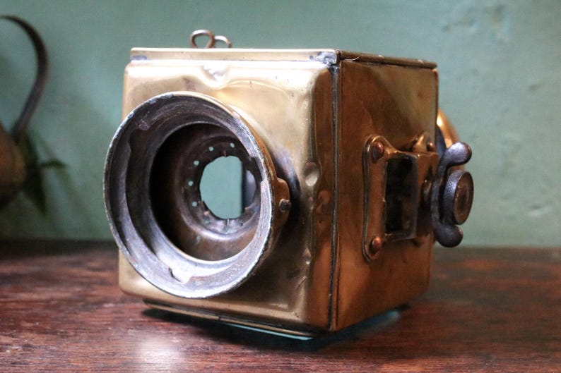 May include: Antique brass camera, square-bodied with a large circular lens. It features a dark metal focusing knob and small rectangular details. The brass has a warm, aged patina, indicating its age. A vintage photographic item.