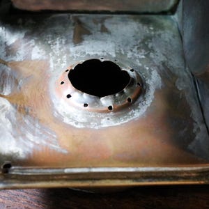 May include: Close-up of the interior of a metal lantern. The base is a mix of copper and silver tones, with a central circular opening surrounded by small holes. The metal shows signs of age and wear.