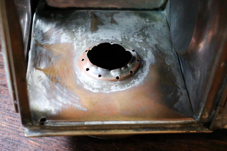 May include: Close-up of the interior of a metal lantern. The base is a mix of copper and silver tones, with a central circular opening surrounded by small holes. The metal shows signs of age and wear.