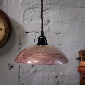 Puede incluir: Lámpara colgante de estilo vintage con una pantalla de cristal moteado de color malva. La pantalla tiene bandas horizontales de color cobre y está suspendida por un cable trenzado marrón. La lámpara tiene una base negra.