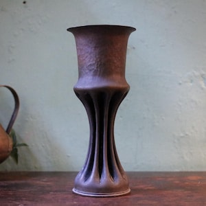 Puede incluir: Un jarrón alto de bronce antiguo con una superficie texturizada y una parte superior acampanada. El jarrón presenta un diseño único con estrías a lo largo de su cuerpo, lo que aumenta su atractivo vintage. El jarrón se exhibe sobre una superficie de madera.