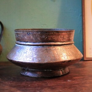 May include: A copper-coloured, antique-style bowl with etched floral and geometric patterns. The bowl has a wide, flared base and a slightly flared rim. The exterior is a warm, reddish-brown colour, and the bowl appears to be made of metal.