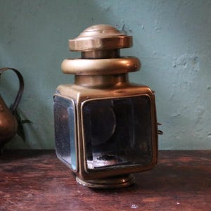 May include: Vintage brass lantern with glass panels. The lantern has a square body with a rounded top and base. The glass panels are clear, and the brass has a warm, aged patina. The lantern is on a wooden surface.