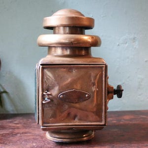 May include: Vintage brass lantern, rectangular with a rounded top. It has a side handle and a glass panel. The brass has a warm, aged patina. A classic lighting fixture, likely from the early 20th century. The lantern is a decorative item.