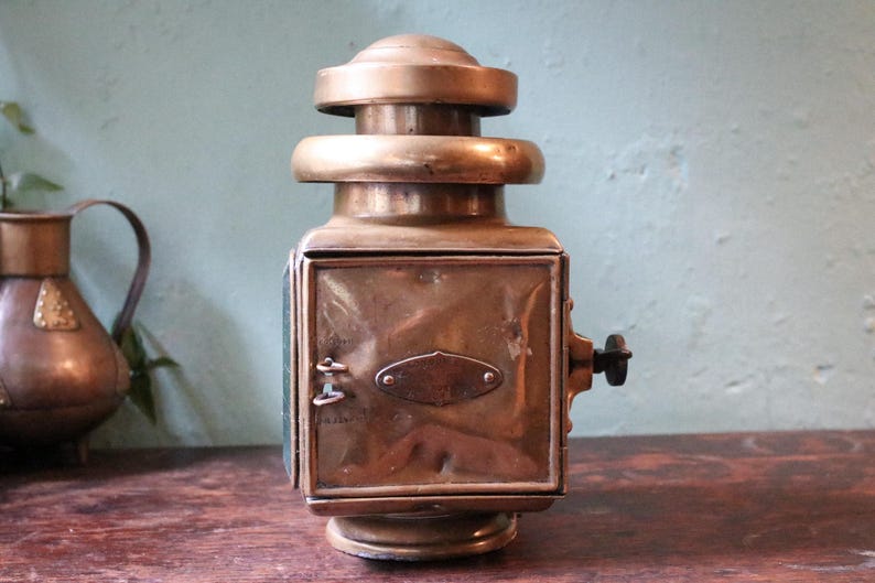 May include: Vintage brass lantern, rectangular with a rounded top. It has a side handle and a glass panel. The brass has a warm, aged patina. A classic lighting fixture, likely from the early 20th century. The lantern is a decorative item.