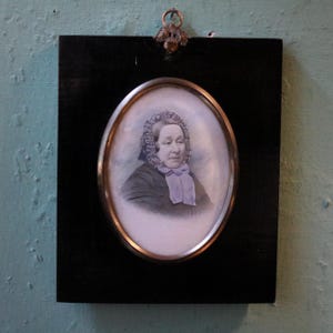 May include: A rectangular black wooden wall plaque featuring an oval portrait of a person wearing a dark bonnet with intricate lace details and a light purple neck scarf. The portrait is encased in a metallic oval frame with a decorative metal hanger.