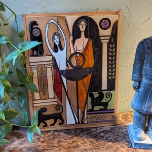May include: A colorful ceramic tile depicting a stylized scene with a woman holding a staff, a bird, and a cat. The tile is framed in wood and has a floral design in the upper right corner.