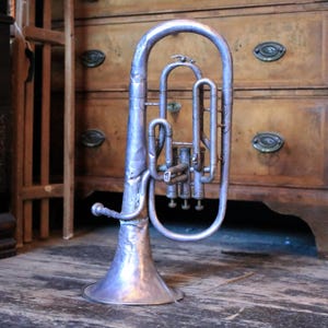 May include: A silver euphonium, a brass instrument, is positioned on a wooden surface. The instrument features a bell, valves, and tubing. A wooden dresser and a wooden structure are in the background.