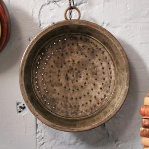 May include: A circular, antique copper colander with many small holes for draining. The colander is suspended by a chain from a ring. The copper has a weathered, aged appearance, suggesting it is vintage. It hangs against a white brick wall.