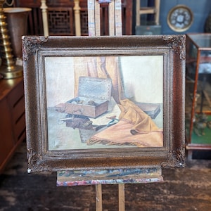 May include: An antique still life painting in a dark brown ornate frame. The painting depicts a wooden box, a yellow cloth, and other objects. The artwork is displayed on a wooden easel in a shop setting.