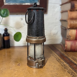 Könnte beinhalten: Eine schwarze und messingfarbene Grubenlampe mit einem Drahtkäfig und einem Griff. Die Lampe hat einen Glaszylinder und einen Messingfuß.