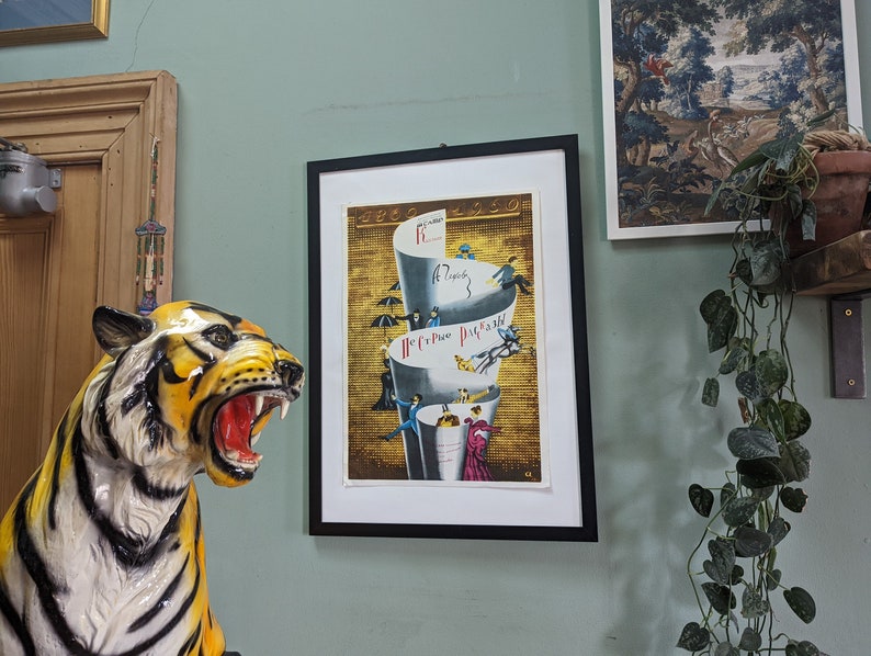 May include: A framed vintage poster with a black border. The poster features a gold background with stacked white cups and small figures. A ceramic tiger statue with yellow and black stripes is in the foreground.