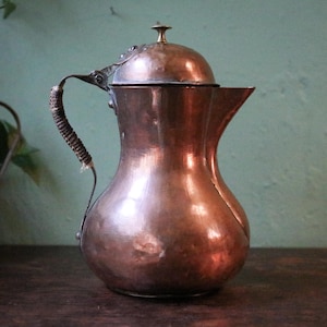 Puede incluir: Una jarra de cobre con tapa y boquilla curva. El asa está envuelta con un material oscuro y texturizado. La jarra tiene un cuerpo bulboso y un pequeño pomo decorativo en la parte superior de la tapa. La jarra es probablemente vintage o antigua.