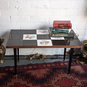 May include: A black and brown tiled coffee table with black metal legs. The table has a floral design on the tiles. There are three books stacked on top of the table, the top book is titled "Plantopedia".