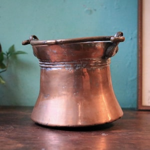 May include: A small, copper-coloured pot with a wide base and a narrow top. It features a handle and a decorative rim. The pot is made of copper and has a shiny surface. It is likely used for cooking or serving. The pot's height is approximately 10 cm.