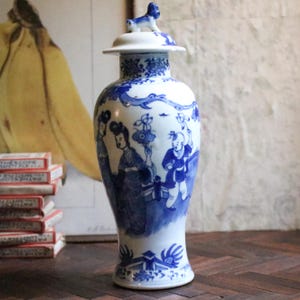 May include: A blue and white porcelain vase with a lid, decorated with hand-painted scenes. The vase depicts figures and floral patterns. A small dog figurine adorns the lid. This decorative vase is a classic piece.