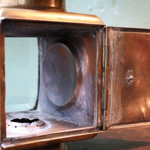 May include: Close-up of a vintage brass lantern with a rectangular body and a hinged door. The lantern features clear glass panels and a circular opening on the side. The door is open, revealing the interior.