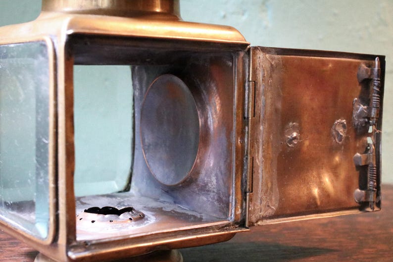 May include: Close-up of a vintage brass lantern with a rectangular body and a hinged door. The lantern features clear glass panels and a circular opening on the side. The door is open, revealing the interior.