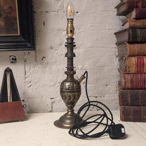 May include: A brass table lamp with an ornate design and a black cord. The lamp is on a white surface and there are books in the background.