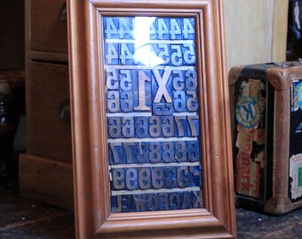May include: A framed collection of vintage letterpress printing blocks. The wooden frame surrounds a grid of individual metal letters, including the letter "X". The blocks are arranged in rows and columns, creating a unique display piece.